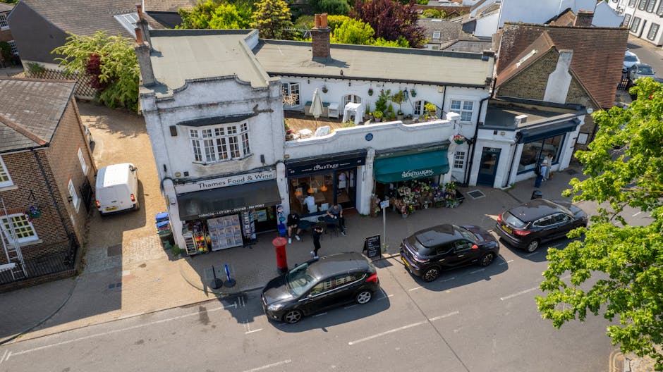 An aerial view of a row of small retail shops on Ruislip Manor High Street, with a white building featuring large display windows and awnings. The pavement in front of the shops is clean, with a red p